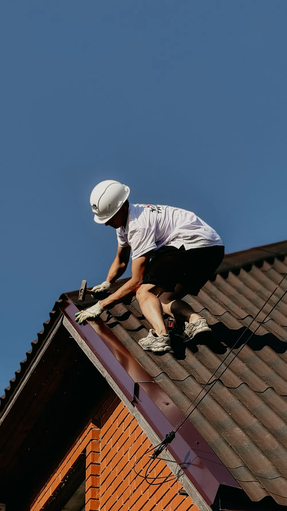 Roofer in a white hard hat nailing flashing along a roof edge under a blue sky